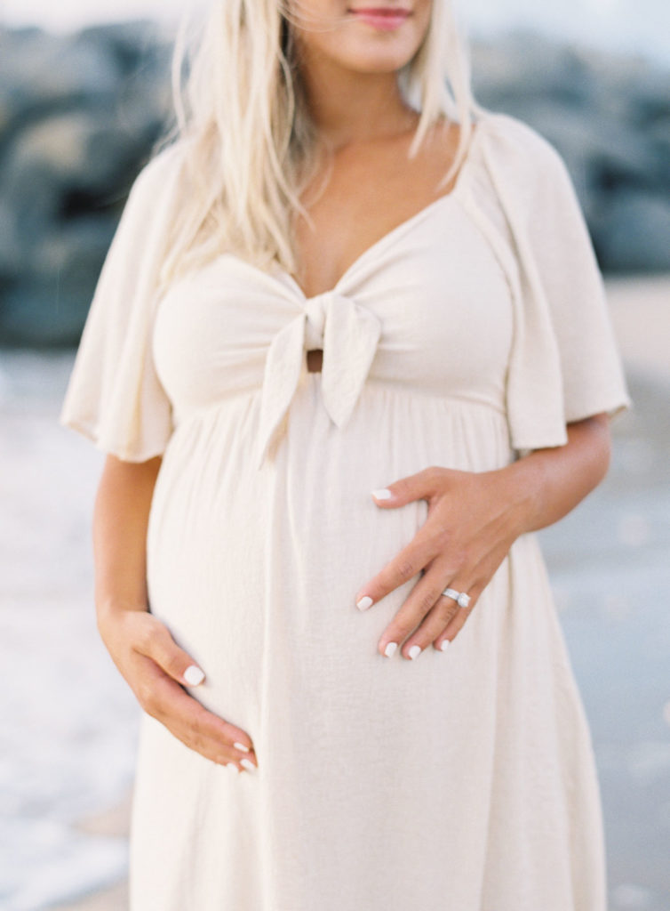 Pregnant women in a cream colored dress holding her belly looking out at the ocean.